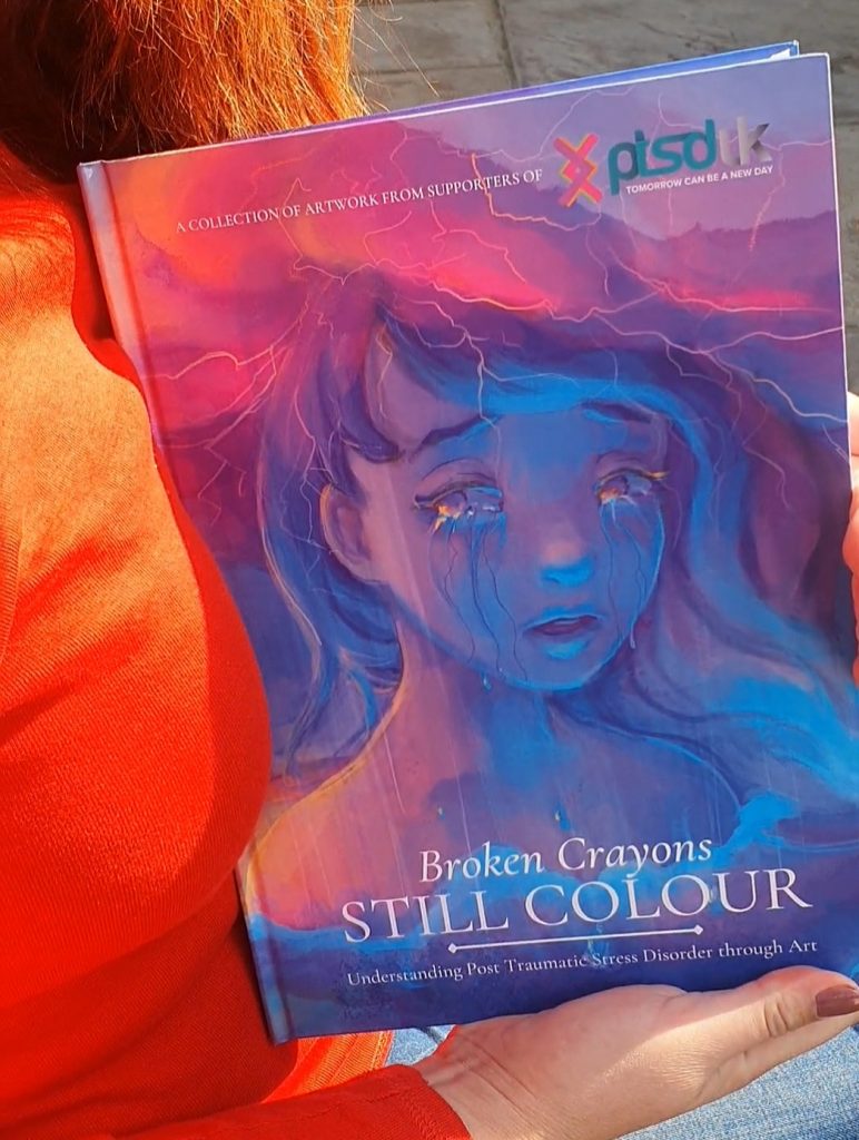 Katy holding a book in her hands, showing the cover of the book 'Broken Crayons Still Colour' how writing can help you to cope with PTSD