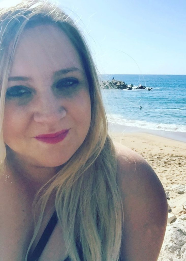 A woman with blond hair on the beach, sea behind her.