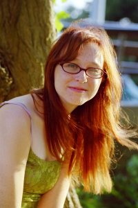 A woman with ginger hair down and glasses, in a green top, in front of a tree