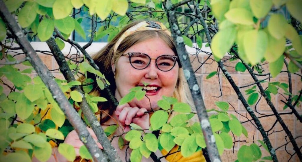 A young lady with glasses and blonde hair is smiling from behind tree branches and green leaves