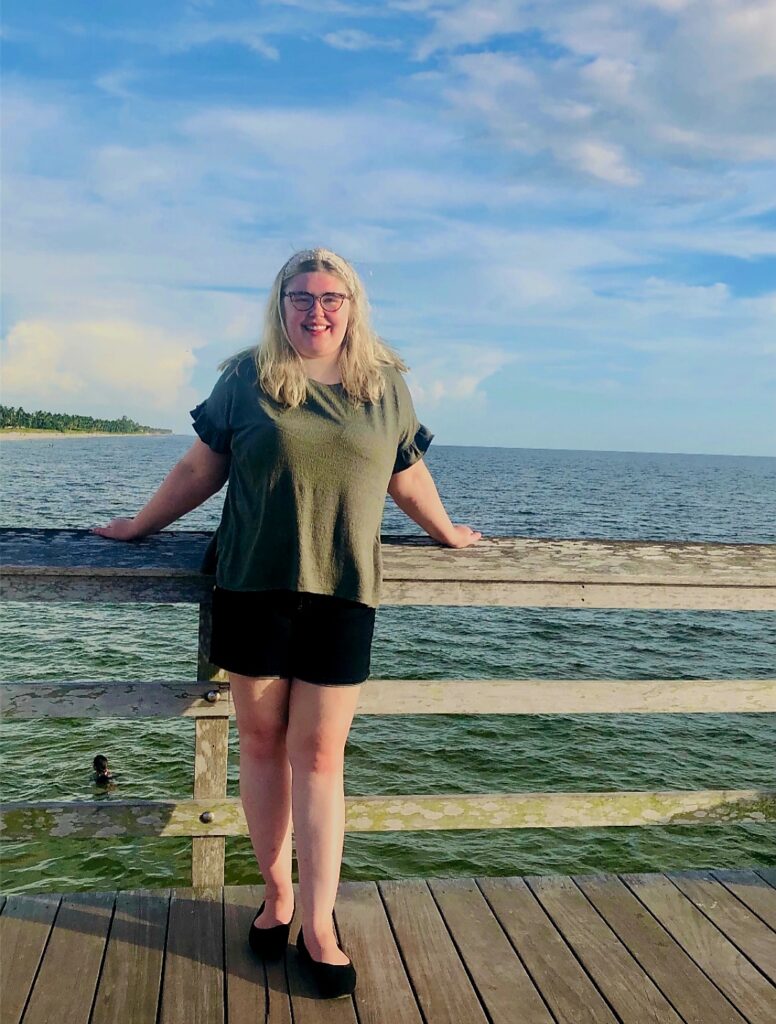 a young lady with blonde hair down and glasses standing in front of the sea