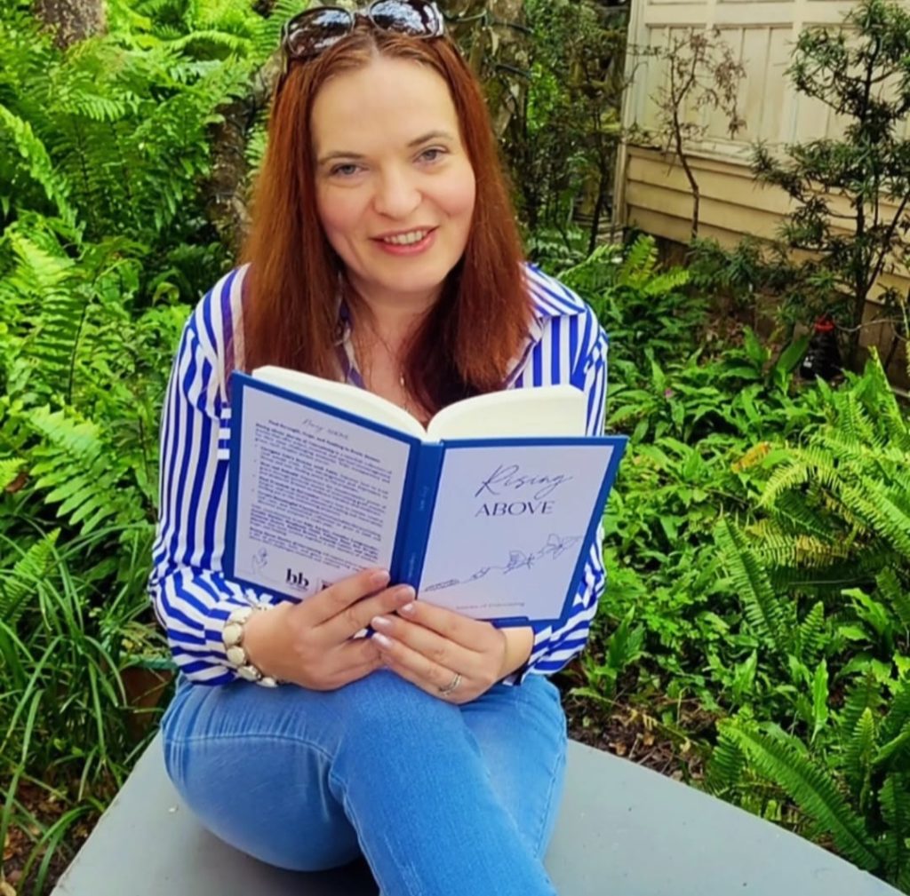 Katy Parker, with brown-reddish hair and a stripy blue and white shirt and blue jeans sitting on a bench in a garden, holding Rising Above book in her hands
